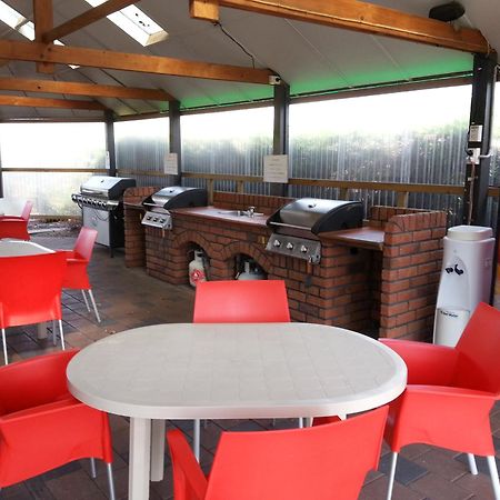 陶波德布雷思汽车旅馆酒店 外观 照片 The photo shows an outdoor kitchen area with a covered space. There are several barbecue grills set up on a brick counter. In the foreground, there are round tables with white tops and bright red chairs arranged around them. A Coca-Cola vending machi