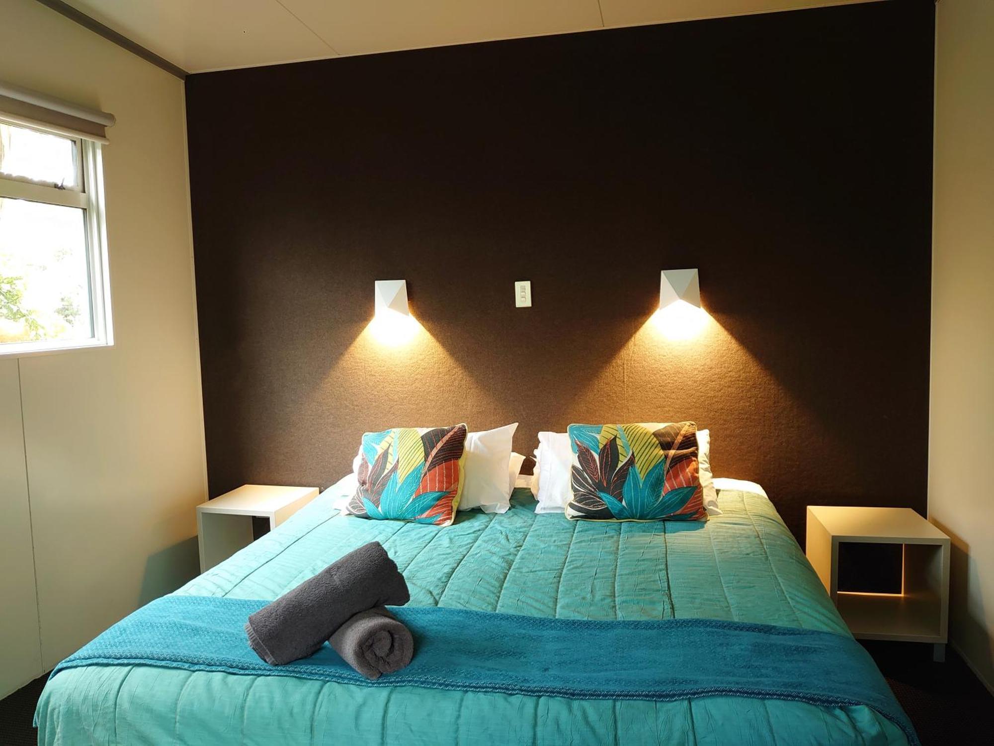 陶波德布雷思汽车旅馆酒店 外观 照片 The image shows a bedroom with a modern and inviting layout. The focal point is a bed with a vibrant turquoise comforter, decorated with two colorful pillows featuring floral patterns. On either side of the bed, there are small nightstands with wall-