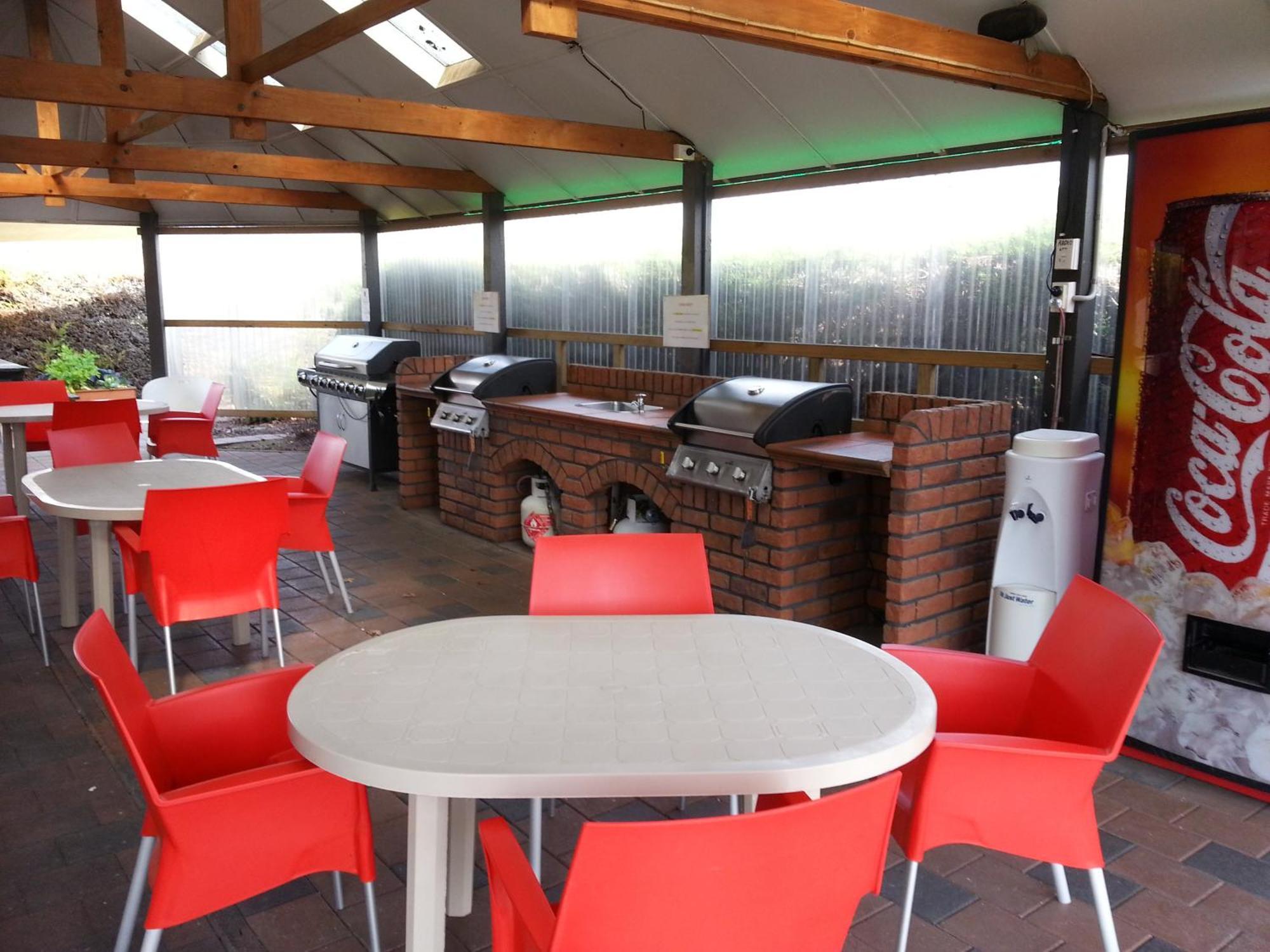 陶波德布雷思汽车旅馆酒店 外观 照片 The photo shows an outdoor kitchen area with a covered space. There are several barbecue grills set up on a brick counter. In the foreground, there are round tables with white tops and bright red chairs arranged around them. A Coca-Cola vending machi